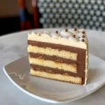 Slice of Salted Caramel cake showing layers of mousse swirls, vanilla cake, and topped with salted caramel whipped creams, and, white and chocolate pearls
