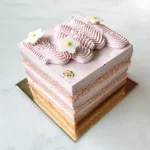 Strawberry Bliss cake from a three-quarter angle — exposed layers of strawberry genoise, namelaka cream and mousse, topped with wavy piped cream, white fondant flowers and gold leaf