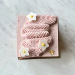 Top-down view of Strawberry Bliss cake — wavy piped strawberry mousse, white fondant flowers with yellow centres, pearl sprinkles and gold leaf.